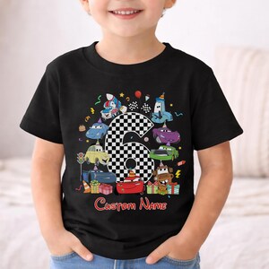 May include: Black t-shirt with a colourful cartoon design of race cars, a chequered number 6, and the text "Custom Name" in red. The design includes birthday-themed elements like balloons and presents. The shirt is for a child.