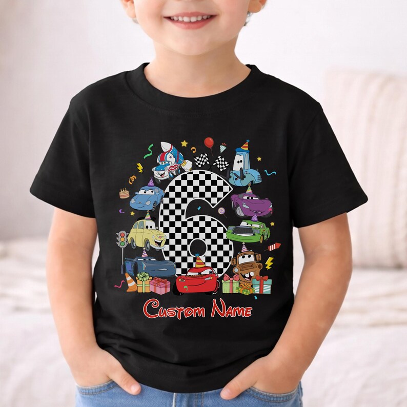 May include: Black t-shirt with a colourful cartoon design of race cars, a chequered number 6, and the text "Custom Name" in red. The design includes birthday-themed elements like balloons and presents. The shirt is for a child.