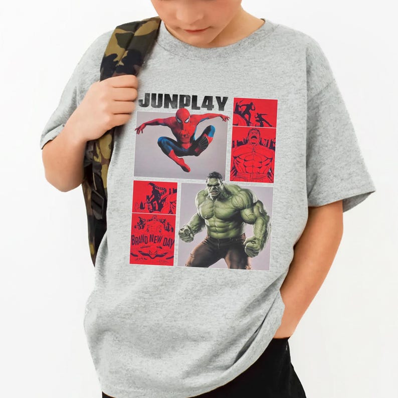 Op de afbeelding: Een grijs t-shirt met een grafisch ontwerp met de tekst "JUNPLAY". Het ontwerp bevat afbeeldingen van Spider-Man en de Hulk, samen met andere stripboekillustraties. De woorden "BRAND NEW DAY" zijn ook zichtbaar.