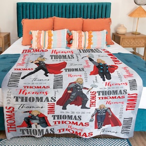 May include: A light gray blanket featuring multiple illustrations of a superhero with a red cape and hammer. The name "Thomas" is printed in red and gray throughout the design. The blanket is on a bed with orange and teal pillows.