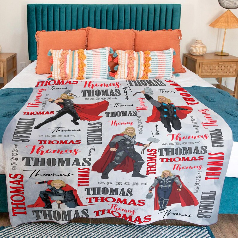 May include: A light gray blanket featuring multiple illustrations of a superhero with a red cape and hammer. The name "Thomas" is printed in red and gray throughout the design. The blanket is on a bed with orange and teal pillows.