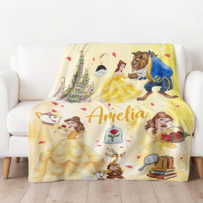 May include: A yellow throw blanket with illustrations of characters from Beauty and the Beast, including Belle and the Beast. The design includes a castle, rose, and other characters. The name "Amelia" is written in gold script.