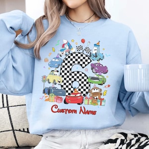 May include: Pale blue sweatshirt with a vibrant print of cartoon cars, a chequered number 6, and the text "Custom Name" in red. The design incorporates birthday elements such as balloons and gifts. A cheerful, celebratory design.