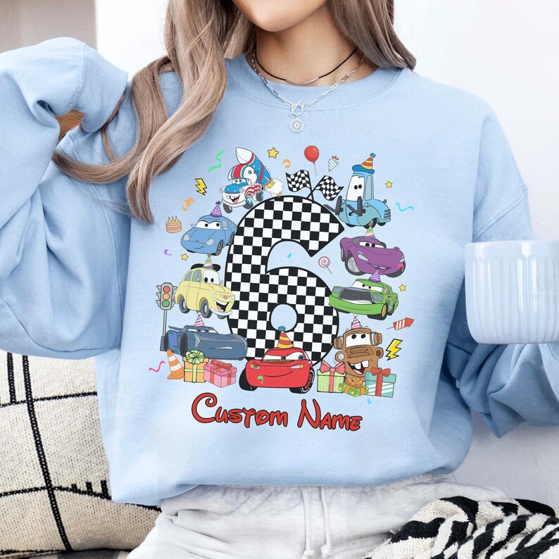 May include: Pale blue sweatshirt with a vibrant print of cartoon cars, a chequered number 6, and the text "Custom Name" in red. The design incorporates birthday elements such as balloons and gifts. A cheerful, celebratory design.