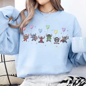 May include: Light blue sweatshirt featuring a graphic of cartoon characters, each holding a Mickey Mouse-shaped balloon. The characters include Stitch, Captain America Stitch, a red Stitch, Hulk Stitch, Wonder Woman Stitch, and Thor Stitch. The design is set against a light blue background.