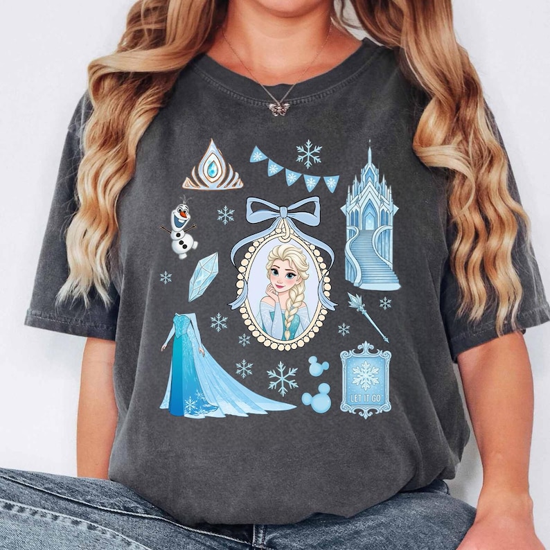 May include: Dark gray t-shirt featuring a collection of blue and white illustrations. The design includes a portrait of a woman with blonde hair, snowflakes, a castle, and the text "LET IT GO".