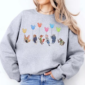 May include: A light gray sweatshirt featuring a colorful graphic of cartoon characters holding Mickey Mouse-shaped balloons. The characters are from the animated movie Zootopia. The balloons are yellow, blue, orange, pink, and green.