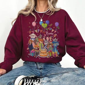 Puede incluir: Sudadera burdeos con un gr&aacute;fico colorido de personajes de dibujos animados, globos y una tarta de cumplea&ntilde;os. El dise&ntilde;o incluye la palabra "Name" y decoraciones festivas, creando un tema de celebraci&oacute;n. La sudadera es de color burdeos liso.