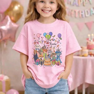 Puede incluir: Camiseta rosa de manga corta con una colorida escena de cumplea&ntilde;os de dibujos animados. El dise&ntilde;o incluye globos, confeti y una tarta. La camiseta tiene la palabra "Name" impresa, y personajes de dibujos animados.