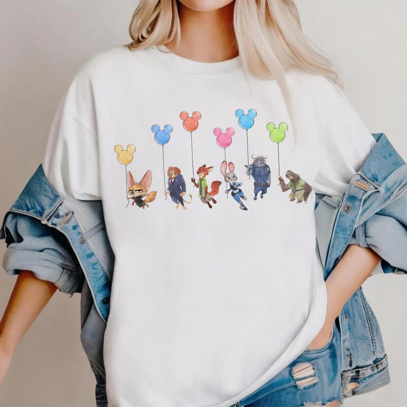 May include: White t-shirt featuring a colorful illustration of characters from the movie Zootopia, each holding a Mickey Mouse-shaped balloon. The balloons are in various colors, including yellow, blue, pink, and green. The design is centered on the shirt.