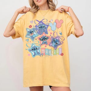 Może przedstawiać: Ż&oacute;łta koszulka z kresk&oacute;wkowym wzorem postaci Stitch wewnątrz gwiazd, ze słowem "magical" w kolorowych literach. Projekt zawiera kokardy, kwiaty i balony. Koszulka jest w stylu casual i wygodnym.