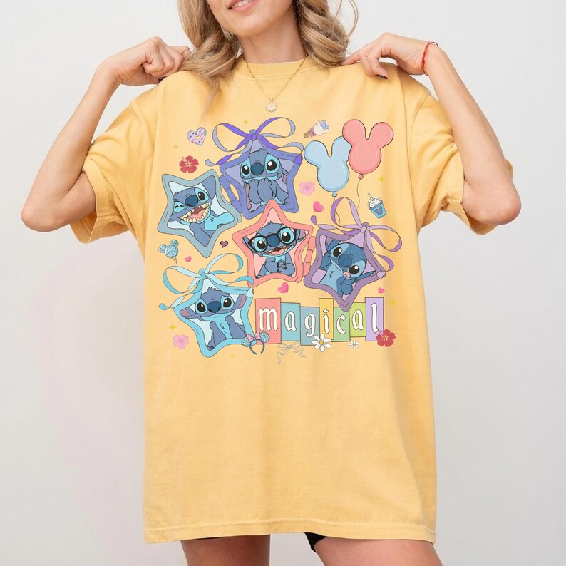 Może przedstawiać: Ż&oacute;łta koszulka z kresk&oacute;wkowym wzorem postaci Stitch wewnątrz gwiazd, ze słowem "magical" w kolorowych literach. Projekt zawiera kokardy, kwiaty i balony. Koszulka jest w stylu casual i wygodnym.