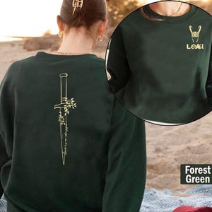 May include: Forest green sweatshirt with a gold graphic of a dagger and floral design on the back. The front of the sweatshirt has a gold graphic of a horned helmet and the word "LOKI".