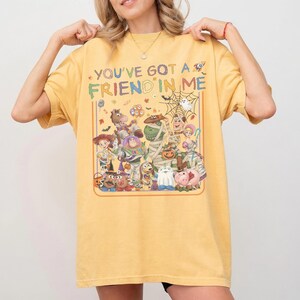 K&ouml;nnte beinhalten: Ein senfgelbes T-Shirt mit einem Grafikdruck verschiedener Zeichentrickfiguren in Halloween-Kost&uuml;men. Der Text "YOU'VE GOT A FRIEND IN ME" ist in bunten Buchstaben &uuml;ber den Figuren gedruckt, darunter eine Mumie, ein Geist und ein K&uuml;rbis.
