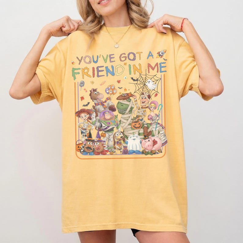 K&ouml;nnte beinhalten: Ein senfgelbes T-Shirt mit einem Grafikdruck verschiedener Zeichentrickfiguren in Halloween-Kost&uuml;men. Der Text "YOU'VE GOT A FRIEND IN ME" ist in bunten Buchstaben &uuml;ber den Figuren gedruckt, darunter eine Mumie, ein Geist und ein K&uuml;rbis.