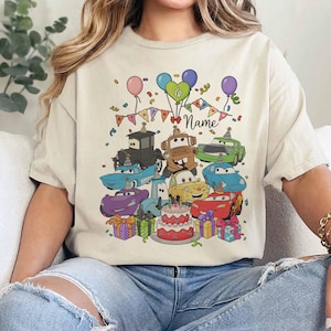 Peut inclure: T-shirt beige avec un motif de voitures de dessins anim&eacute;s color&eacute;s, des ballons, des confettis et un g&acirc;teau d'anniversaire. Le motif comprend le mot "Name" sur une banni&egrave;re, parfait pour un cadeau d'anniversaire personnalis&eacute;.