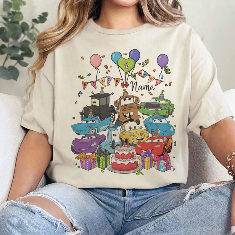 Peut inclure: T-shirt beige avec un motif de voitures de dessins anim&eacute;s color&eacute;s, des ballons, des confettis et un g&acirc;teau d'anniversaire. Le motif comprend le mot "Name" sur une banni&egrave;re, parfait pour un cadeau d'anniversaire personnalis&eacute;.