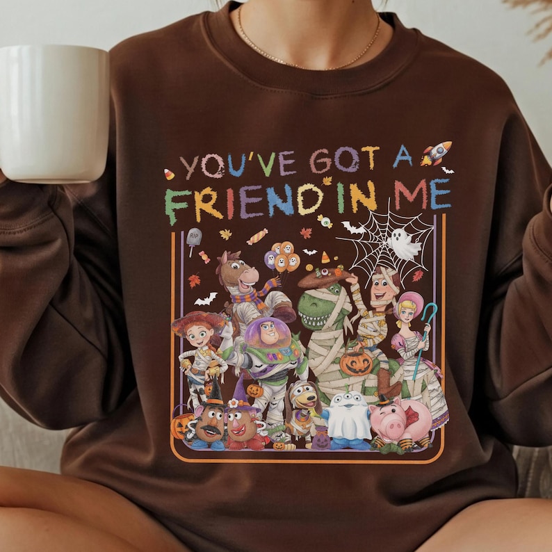 K&ouml;nnte beinhalten: Ein dunkelbraunes Sweatshirt mit Rundhalsausschnitt und dem Schriftzug "YOU'VE GOT A FRIEND IN ME" in bunten Buchstaben &uuml;ber einer Grafik verschiedener Zeichentrickfiguren in Halloween-Kost&uuml;men, darunter eine Mumie, ein Geist und K&uuml;rbisse.