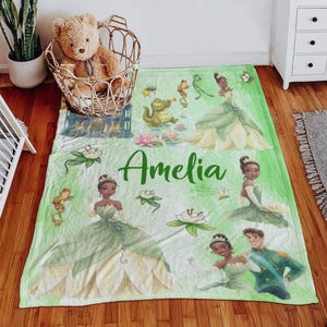 May include: A light green blanket with illustrations of a princess, a frog, and a prince. The blanket has the name "Amelia" in green script. The design includes water lilies and a cartoon alligator playing a trumpet.