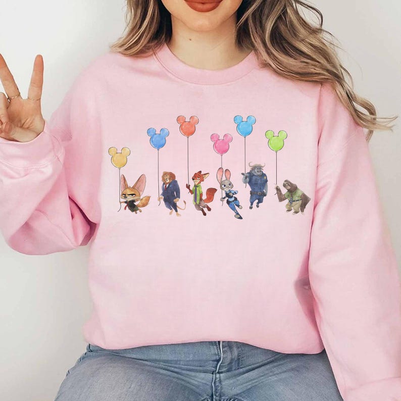 May include: A light pink sweatshirt featuring a colorful graphic of cartoon characters holding Mickey Mouse-shaped balloons. The characters are from the movie Zootopia. The balloons are yellow, blue, pink, and green.