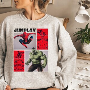 Op de afbeelding: Een lichtgrijs sweatshirt met een grafisch ontwerp. Het ontwerp bevat de tekst "JUNPLAY", Spider-Man en Hulk, met rode en zwarte accenten. De woorden "Brand New Day" zijn ook zichtbaar.