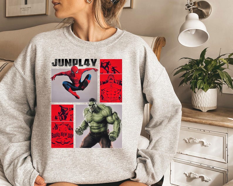 Op de afbeelding: Een lichtgrijs sweatshirt met een grafisch ontwerp. Het ontwerp bevat de tekst "JUNPLAY", Spider-Man en Hulk, met rode en zwarte accenten. De woorden "Brand New Day" zijn ook zichtbaar.