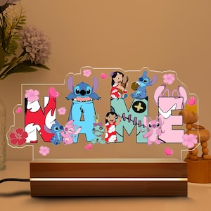 May include: A decorative night light with the word "NAME" and cartoon characters. The letters are in blue, pink, and red, with floral and heart accents. The light sits on a wooden base.