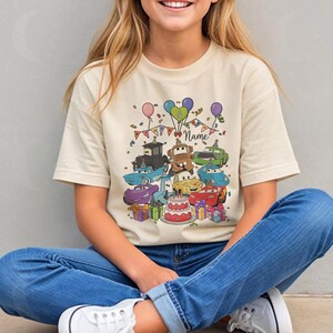 Peut inclure: T-shirt beige avec un motif color&eacute; de voitures de dessins anim&eacute;s, un g&acirc;teau d'anniversaire, des ballons et des confettis. Le motif comprend le mot "Name" et est entour&eacute; de bo&icirc;tes cadeaux. Le t-shirt est port&eacute; par une personne portant un jean bleu.
