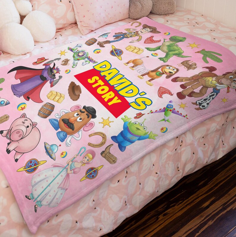Puede incluir: Una manta de forro polar rosa claro con un patr&oacute;n repetitivo de personajes de Toy Story, incluyendo a Woody, Buzz Lightyear, Rex, Slinky Dog, Bullseye, Mr. Potato Head, Hamm y Bo Peep. Un rect&aacute;ngulo rojo central muestra el texto DAVID'S STORY.