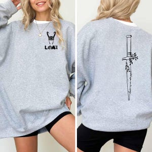 May include: A light gray sweatshirt with a white collar. The front features a black graphic of Loki's helmet and the name "LOKI". The back displays a black dagger design with the words "Worth a Strong".