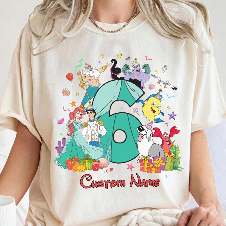 K&ouml;nnte beinhalten: Ein cremefarbenes T-Shirt mit einer gro&szlig;en t&uuml;rkisfarbenen Zahl sechs, umgeben von Zeichentrickfiguren wie einer Meerjungfrau, einem Prinzen, einer Meerhexe, einem gelben Fisch, einem Krebs und einem Hund, die alle Partyh&uuml;te tragen.