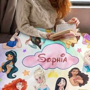 May include: A personalized blanket featuring watercolor illustrations of Disney princesses. The blanket has a light blue and white background with the name "Sophia" in a pink cloud. Includes images of Snow White, Jasmine, Elsa, and others.