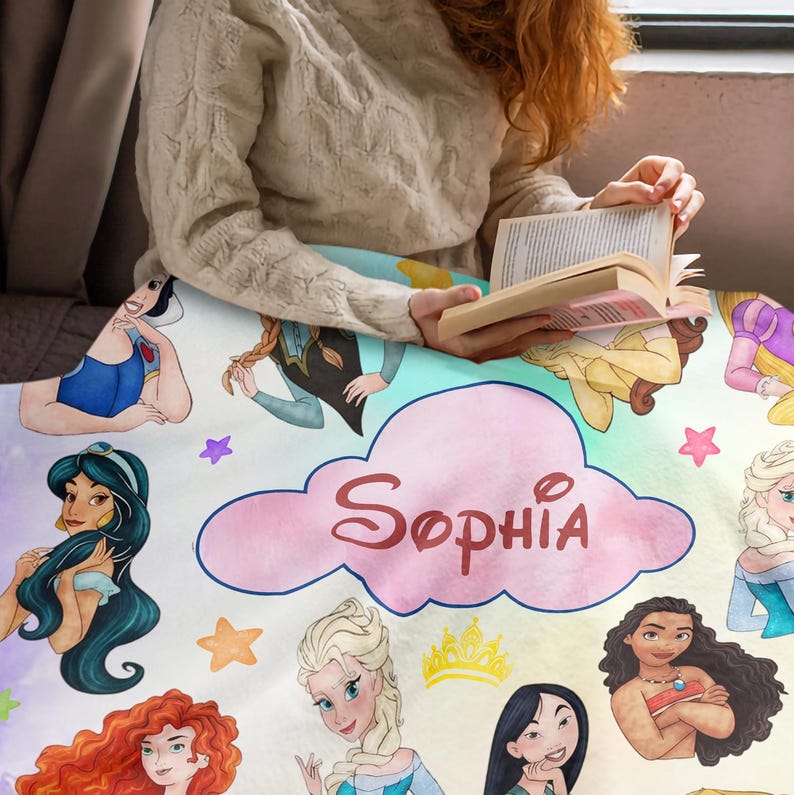 May include: A personalized blanket featuring watercolor illustrations of Disney princesses. The blanket has a light blue and white background with the name "Sophia" in a pink cloud. Includes images of Snow White, Jasmine, Elsa, and others.