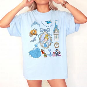 May include: Light blue t-shirt featuring a collection of Cinderella-themed illustrations. The design includes a portrait of Cinderella, a pumpkin carriage, a glass slipper, a blue dress, and other related elements. The shirt is a comfortable, casual style.