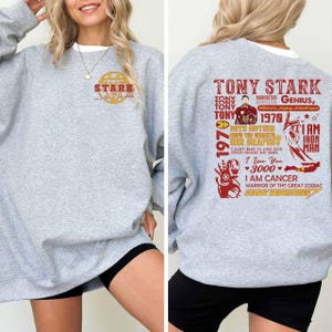 May include: A light gray sweatshirt with a white crew neck collar. The front features a gold and red circular design with the word "STARK". The back displays a red graphic with the words "TONY STARK" and other text, including "IRON MAN".