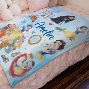 May include: A light blue blanket with a watercolour illustration of Snow White and other characters. The blanket features the name "Amelia" in cursive script, along with images of the Evil Queen, the dwarfs, and a castle. The blanket is suitable for a child.