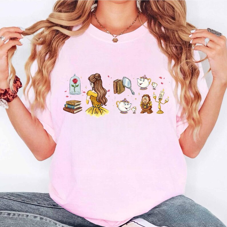 Peut inclure: T-shirt rose clair avec une illustration de dessin anim&eacute; d'une princesse en robe jaune, d'une rose dans une cloche, de livres, de tasses &agrave; th&eacute; et d'autres personnages. Le design est de style dessin anim&eacute;.