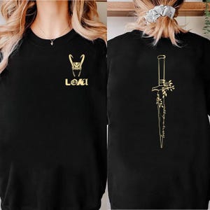 May include: Black sweatshirt featuring a gold-colored Loki logo and text. The back of the sweatshirt displays a gold-colored dagger design with floral accents and script. A casual, comfortable garment.