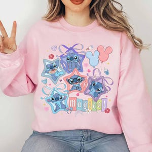 Może przedstawiać: R&oacute;żowa bluza z nadrukiem Stitcha z Lilo & Stitch w ramkach w kształcie gwiazd. Słowo "magical" jest wydrukowane pod postaciami. Projekt zawiera balony, kwiaty i kokardy.