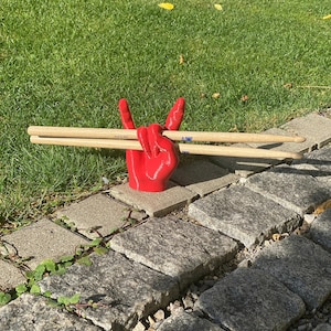 May include: A red hand-shaped drumstick holder in a peace sign gesture, holding two wooden drumsticks. The holder is on a stone walkway with green grass in the background.