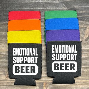 May include: Two black neoprene can coolers with the text "Emotional Support Beer" in white. The coolers are stacked on top of colorful neoprene sleeves in red, orange, yellow, green, blue, and purple.