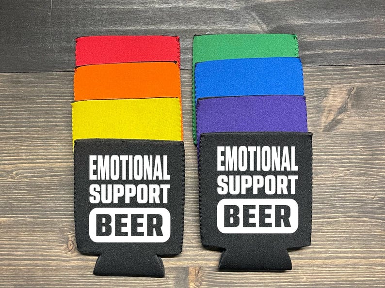 May include: Two black neoprene can coolers with the text "Emotional Support Beer" in white. The coolers are stacked on top of colorful neoprene sleeves in red, orange, yellow, green, blue, and purple.