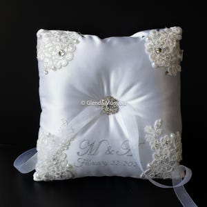 May include: White satin ring pillow with lace and pearl embellishments. Features a silver brooch and ribbon. The pillow has the initials "M & S" and the date "February 22, 2021" embroidered in silver. A decorative item for a wedding.