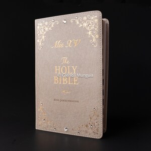 May include: A beige leather-bound Bible with gold lettering and floral embellishments. The cover features the text "Mis XV" and "The Holy Bible, King James Version." Rhinestones are set into the corners. The copyright notice "© Glenda Munguia" is also visible.