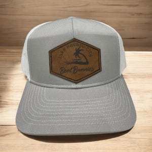 Puede incluir: Una gorra de camionero gris y blanca con un parche de cuero marrón que dice "Lake Havasu City Boat Bunnies".