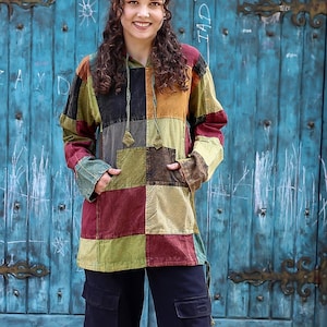 Può includere: Una camicia con cappuccio a patchwork con maniche lunghe e vestibilità ampia. La camicia è realizzata con tessuti di diversi colori, tra cui rosso, verde, giallo e marrone. La modella indossa la camicia con pantaloni cargo neri.