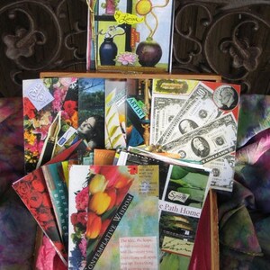May include: A wooden box containing a collection of handmade art cards. The cards showcase collages with floral, botanical, and abstract designs. Some cards include text, and one displays US currency. The box rests on a colourful fabric.