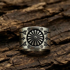 May include: A wide silver ring featuring a central sunburst or flower motif surrounded by engraved arrow designs pointing outward. The metal has a polished finish with darkened recessed areas to highlight the intricate geometric patterns.