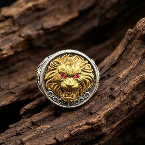 Può includere: Un anello circolare con una testa di leone color oro, una criniera dettagliata e due piccole pietre rosse come occhi. Il leone è centrato su uno sfondo scuro e strutturato all'interno di una cornice color argento lucido con decorazioni a volute.