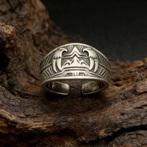 Puede incluir: Un anillo plateado con un diseño grabado que presenta dos águilas flanqueando un motivo central de montaña. El anillo tiene un diseño abierto y está sobre un fondo oscuro y texturizado.